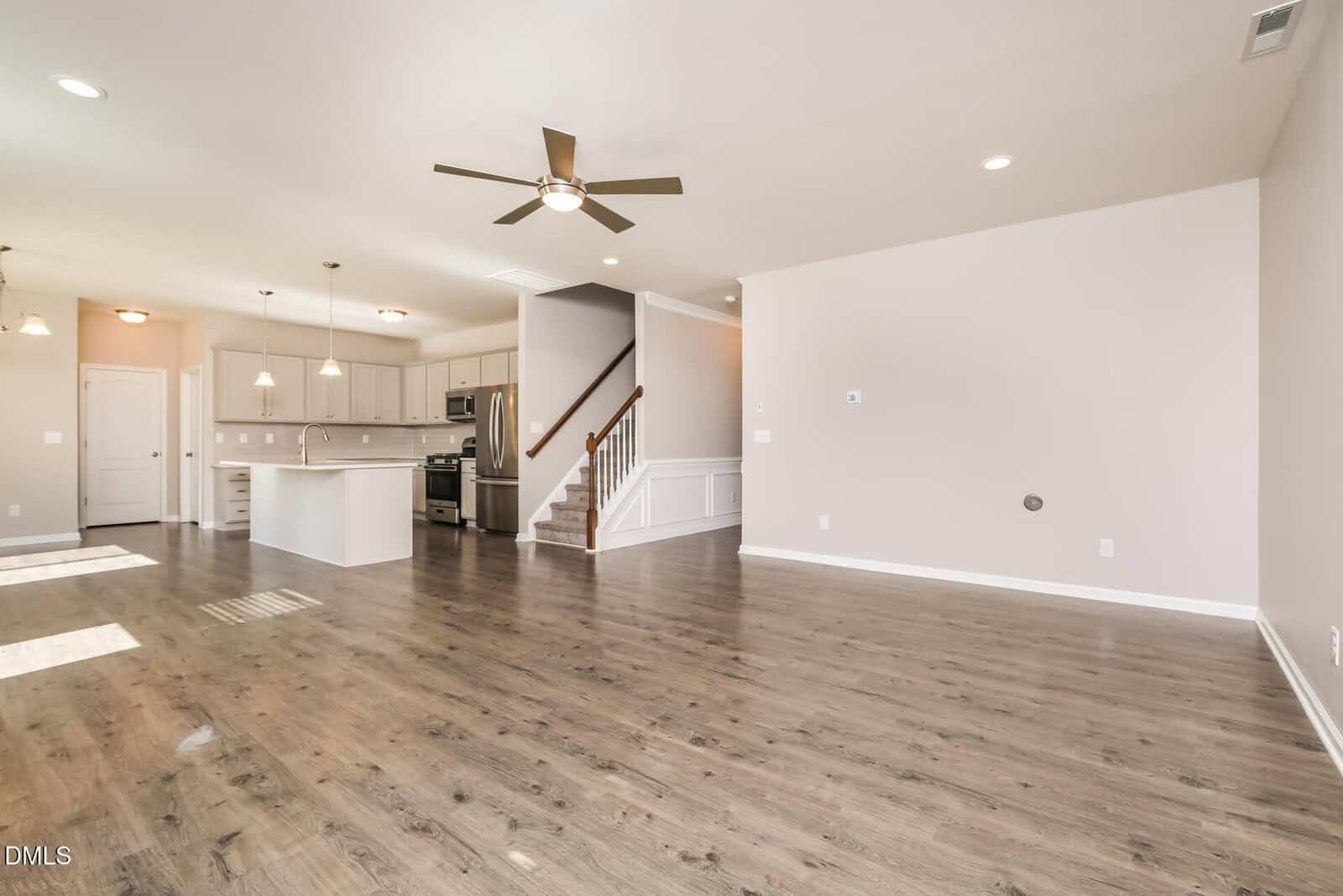 Open-concept living space with wood flooring, neutral walls, a ceiling fan, recessed lighting, a modern kitchen with white cabinets and an island, and a staircase leading upstairs. Natural light comes from windows on the left.