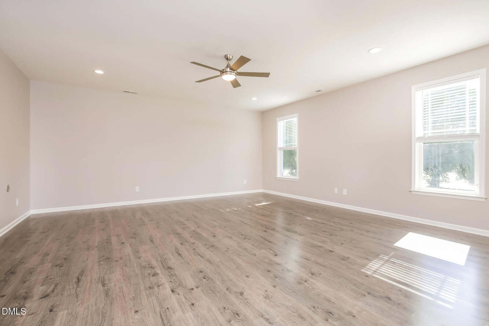 A spacious, empty room with light-colored walls, wood-style flooring, two windows letting in natural light, recessed ceiling lights, and a ceiling fan in the center.