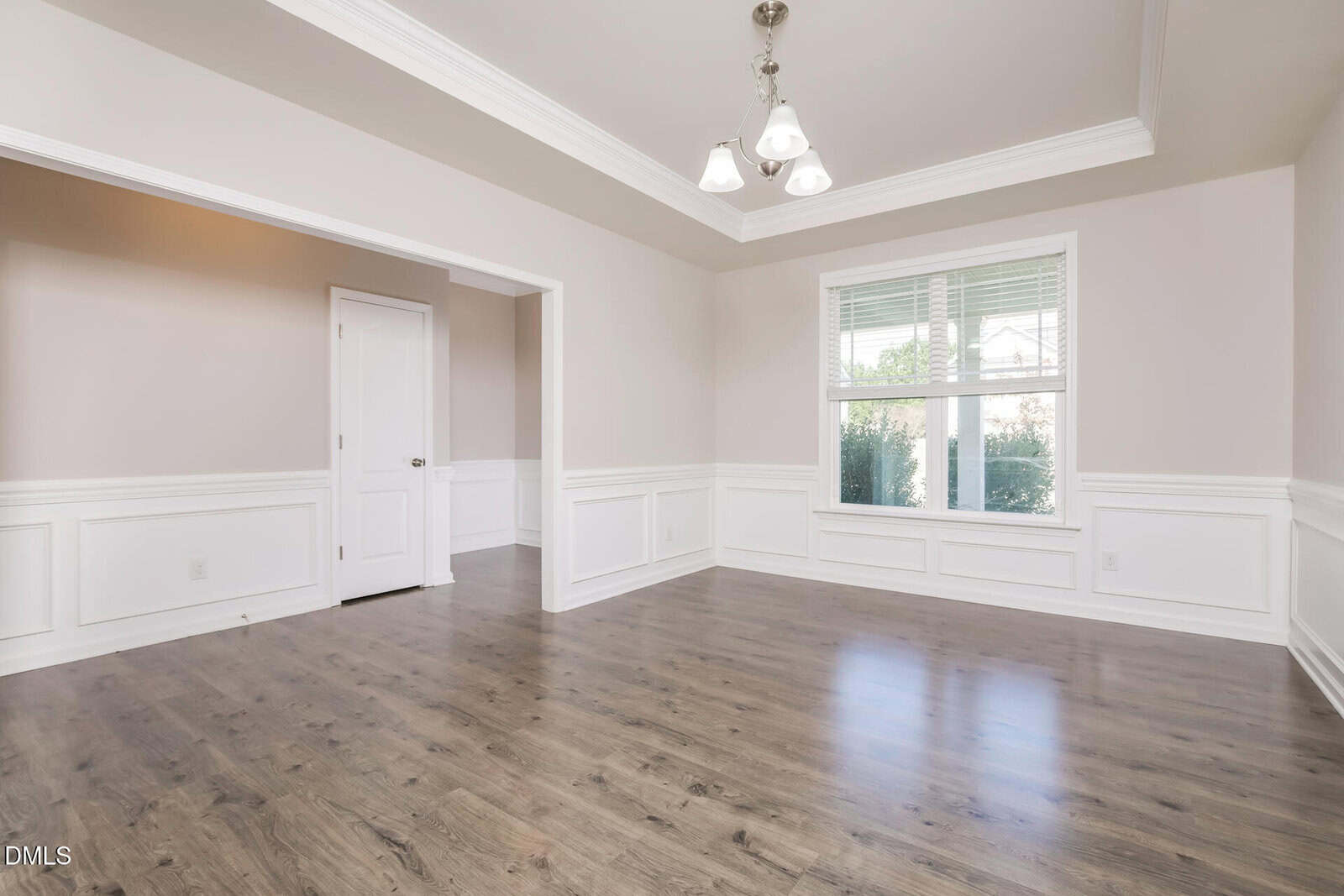 A bright, empty room with light gray walls, white wainscoting, wood-look flooring, a large window with blinds, and a central ceiling light fixture. A white door is visible to the left.