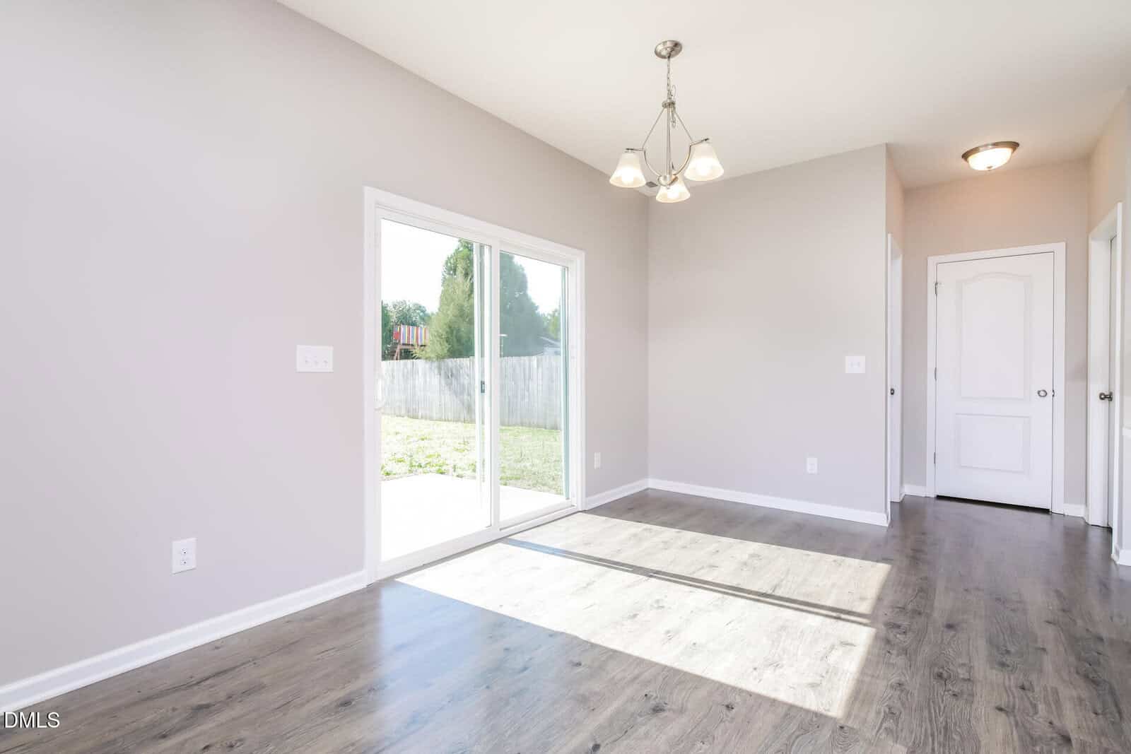 A bright, empty room with light gray walls, wood-style flooring, a ceiling light fixture, sliding glass doors to a fenced backyard, and a hallway leading to a closed white door.