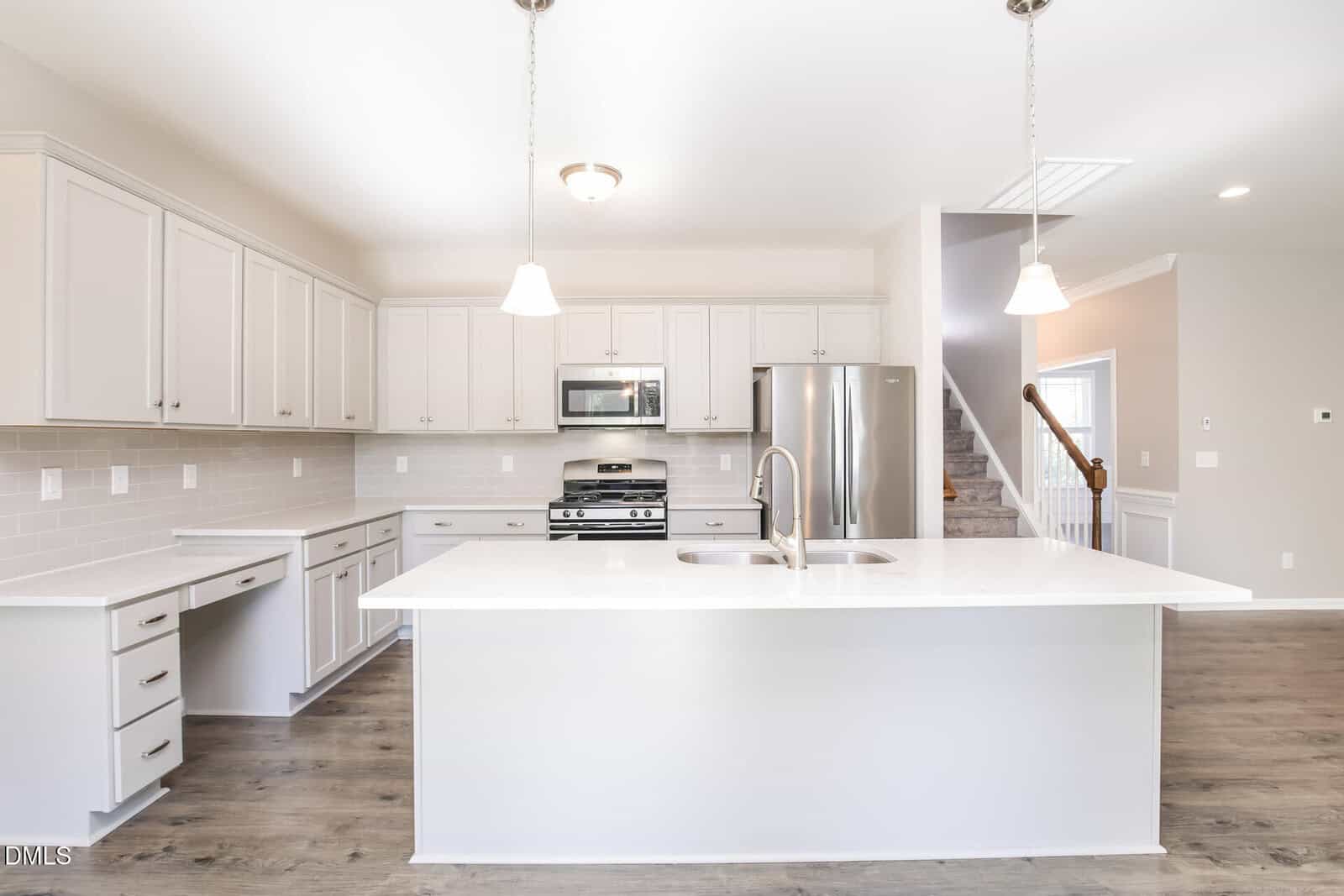 Bright modern kitchen with white cabinetry, stainless steel appliances, large island with sink, pendant lighting, and light wood flooring. Stairs and open entryway are visible in the background.