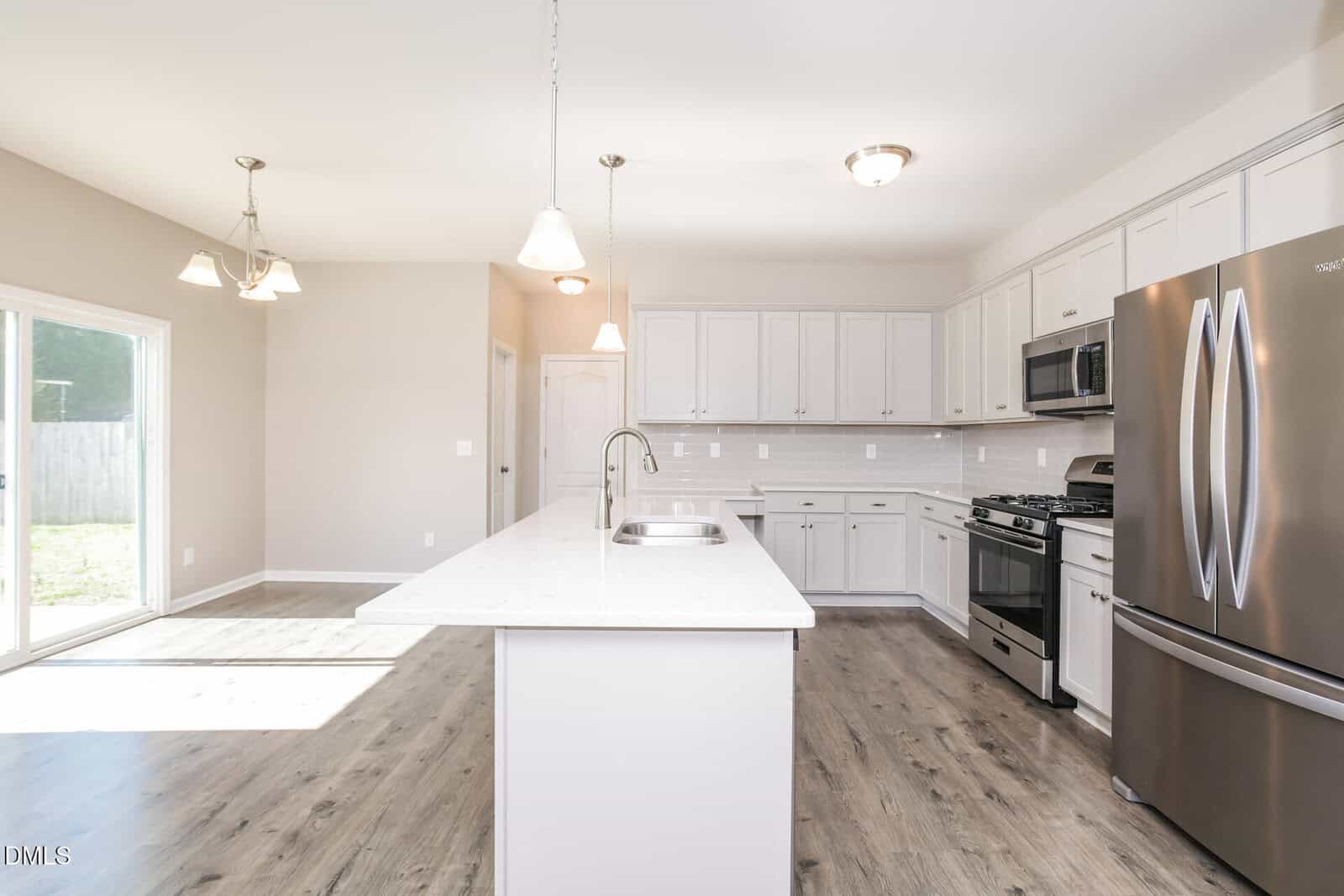Modern kitchen with white cabinets, stainless steel appliances, a large island with sink, pendant lighting, and light wood flooring. Sliding glass doors let in natural light from the left.