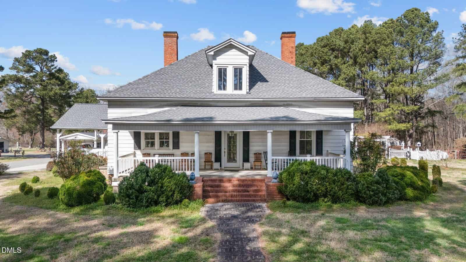 A white, single-story house with a gray roof, two brick chimneys, a small central upper window, and a covered front porch, surrounded by bushes and trees on a sunny day.