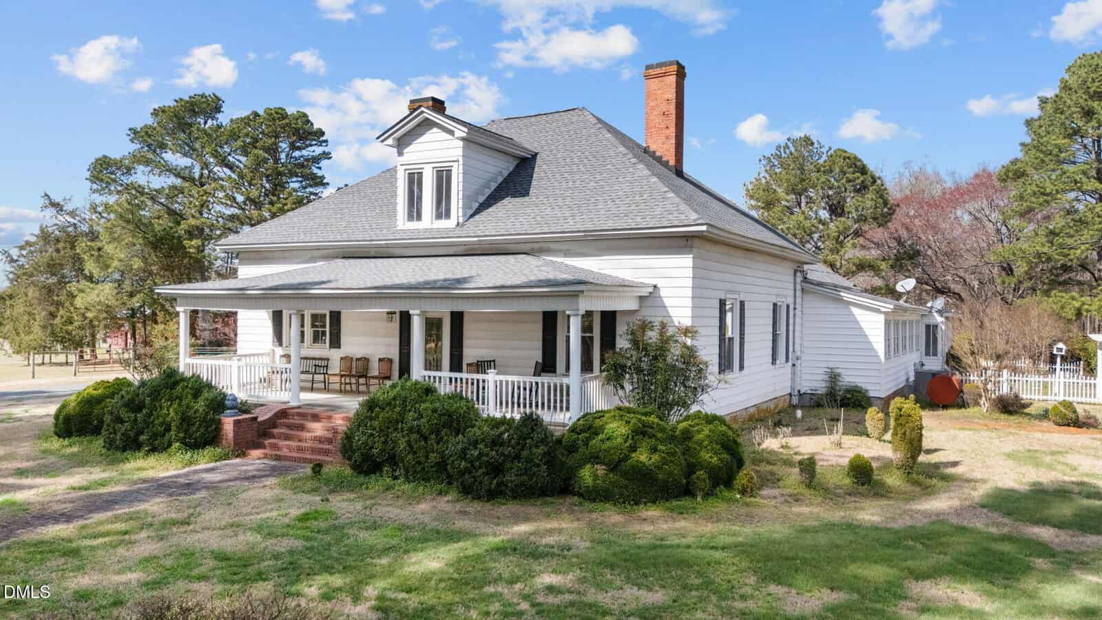 A white, single-story house with a gray roof, front porch with chairs, red brick steps, and surrounding shrubs. The house is set on a grassy lot with trees in the background under a blue sky with clouds.