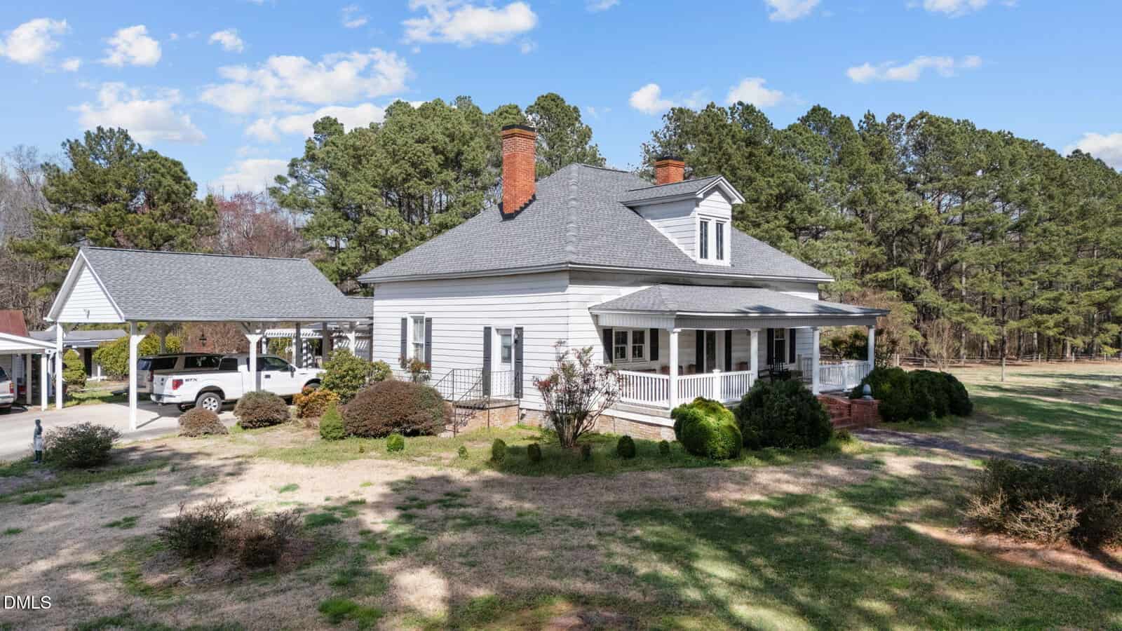 A white, single-story house with a front porch and gabled roof sits on a grassy lot with shrubs and trees. A detached carport with two white vehicles is to the left. Tall pine trees line the background under a blue sky.