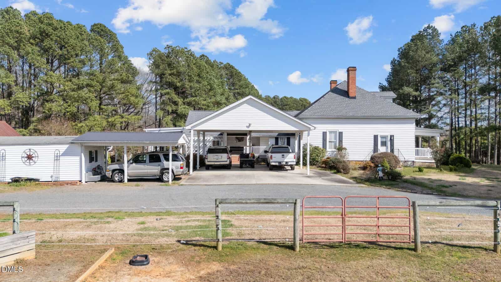 A white farmhouse with a covered driveway and two vehicles parked underneath, flanked by trees on both sides. A wooden fence and red metal gate are in the foreground, with clear skies overhead.