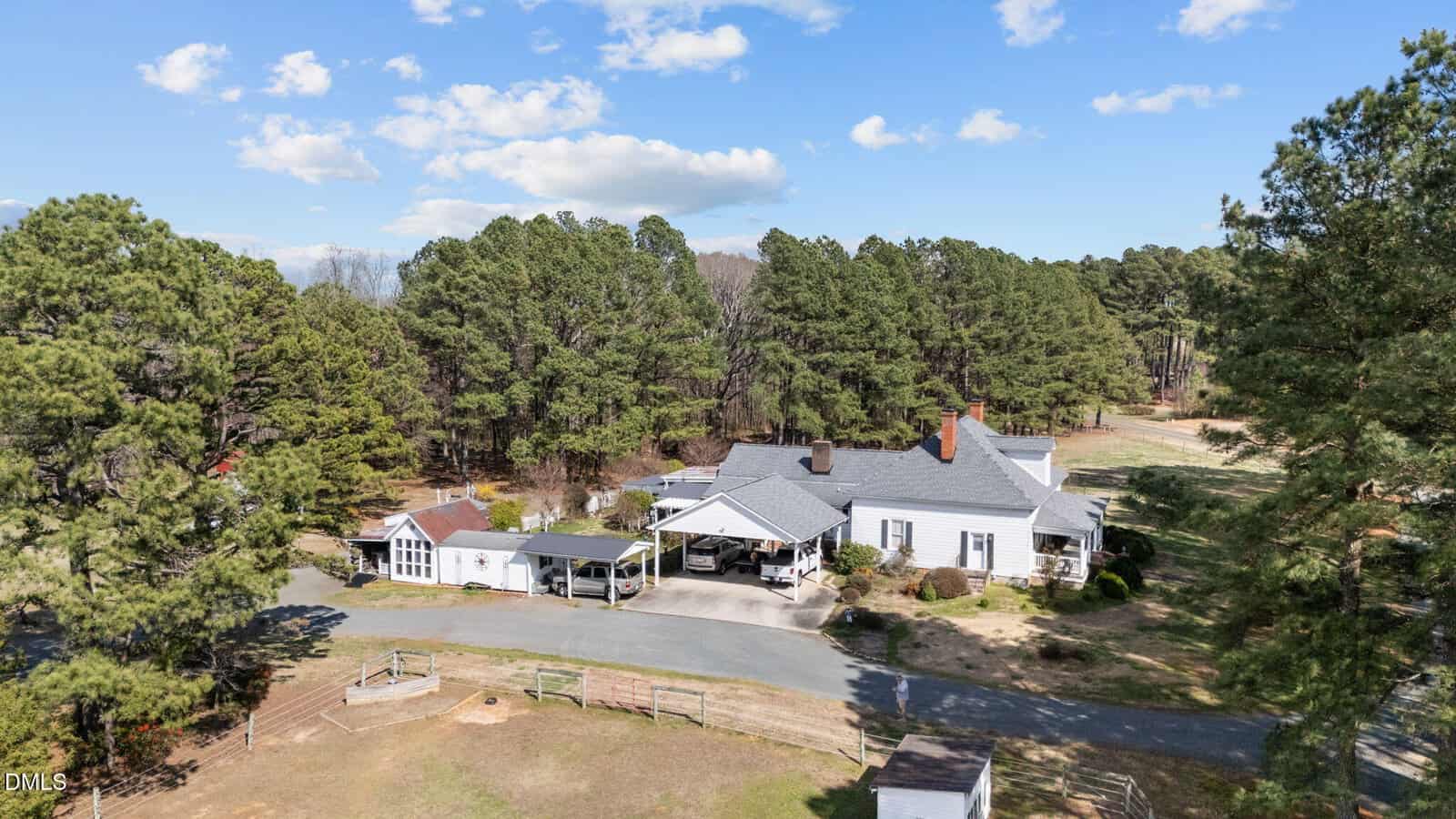 Aerial view of a white house with a gray roof, surrounded by tall trees and a dirt driveway. The property includes a small fenced area, outbuildings, and open space under a partly cloudy sky.