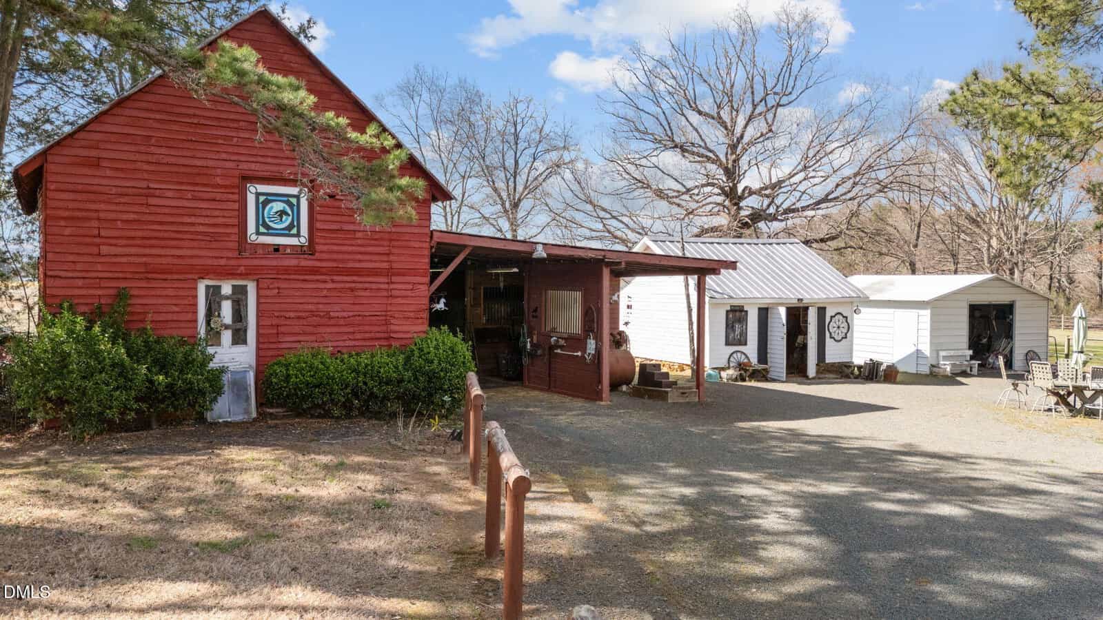A red wooden barn with a sign on the front sits next to two smaller white buildings on a gravel lot. There are some outdoor tables, leafless trees, green bushes, and a partly cloudy sky in the background.
