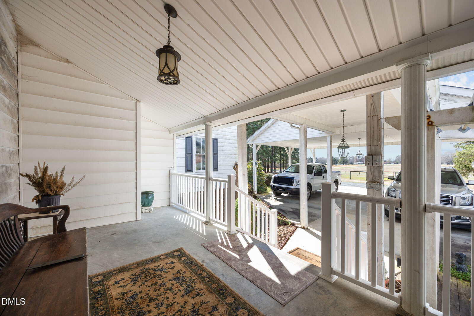 A covered front porch with a sloped ceiling, white railing, and a hanging light fixture. There is a bench, a rug, and a potted plant. Two cars are parked in the driveway visible beyond the porch.