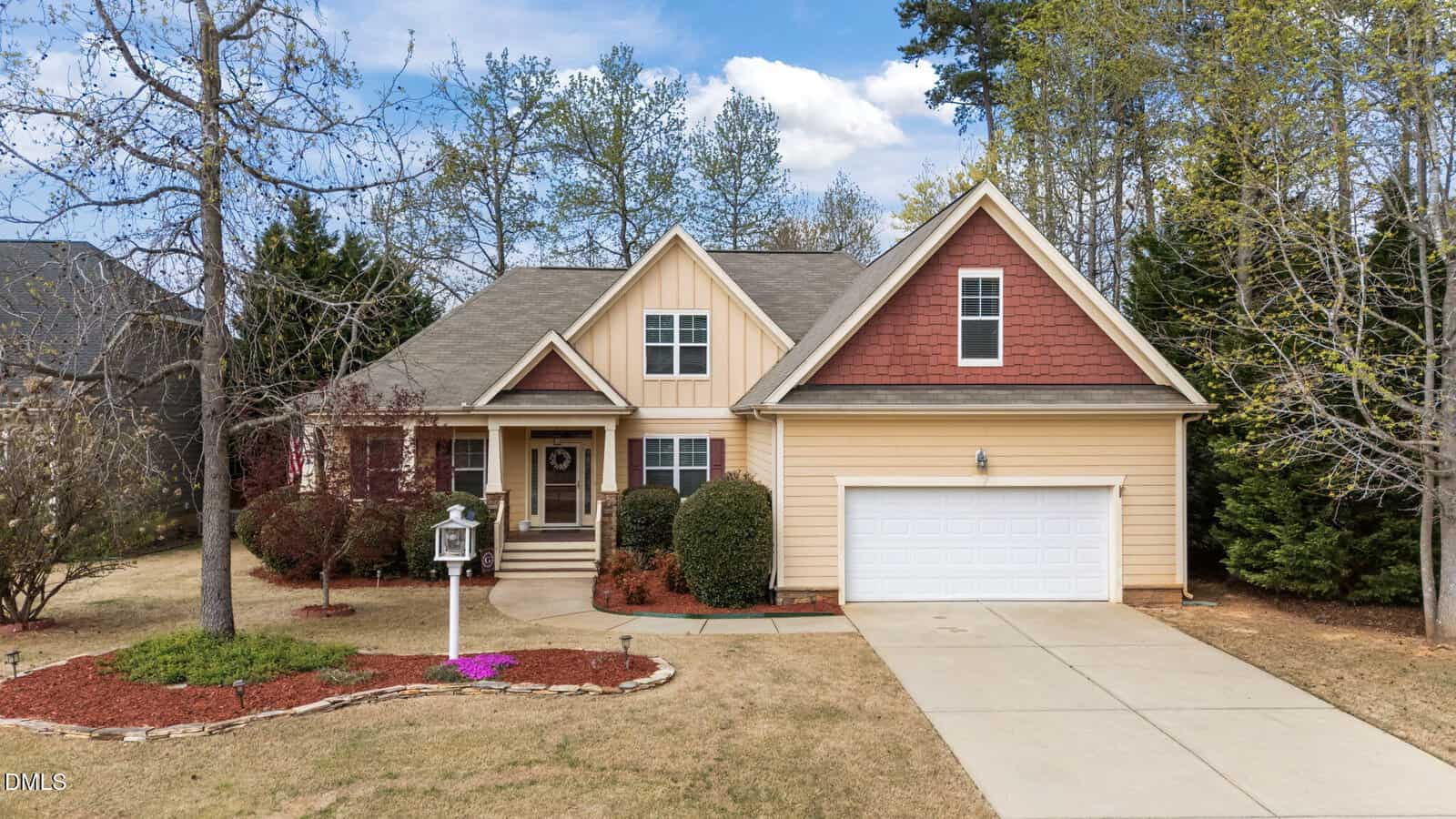 A beige and red suburban house with a two-car garage, a driveway, small front porch, landscaped yard with mulch, shrubs, and trees, under a partly cloudy sky.