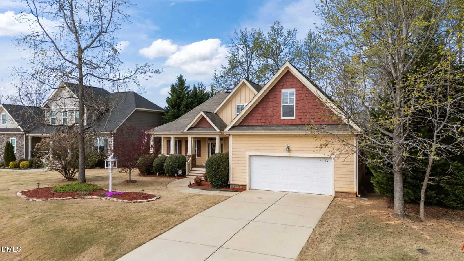 A beige two-story house with white trim, a front porch, attached two-car garage, gabled roof, and a well-maintained front yard with trees and shrubs, situated in a suburban neighborhood.
