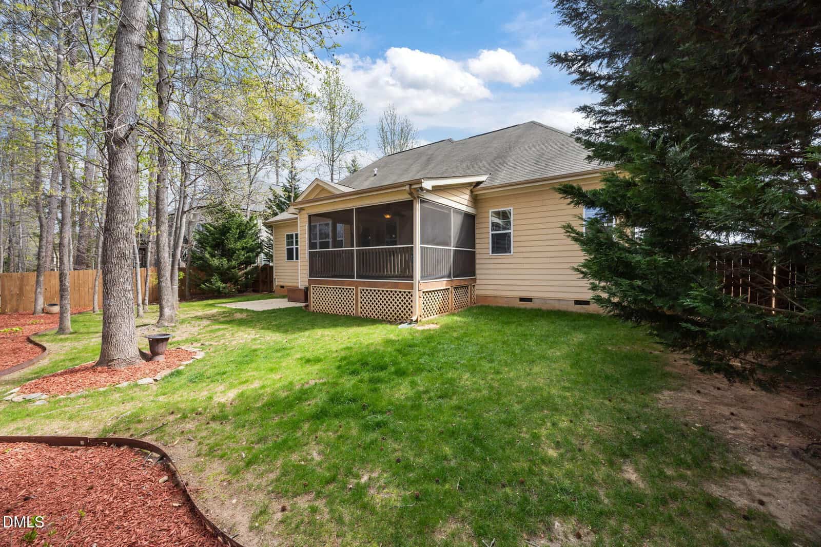 A beige house with a screened-in porch sits in a grassy backyard bordered by mulch, trees, and a wooden fence, under a partly cloudy sky.