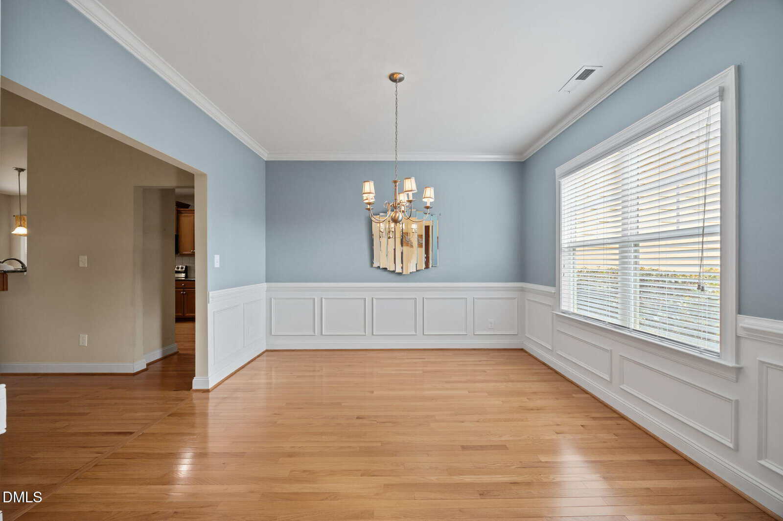 A bright, empty dining room with light blue and white walls, wooden floors, a gold chandelier, and a large window with white blinds letting in natural light. An open doorway leads to another room.