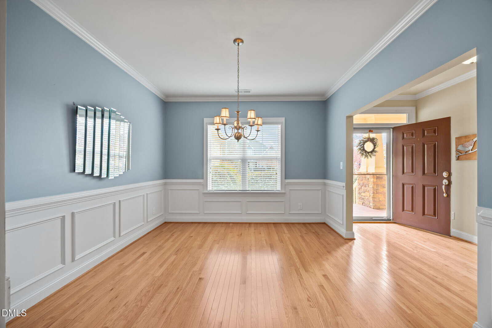 A spacious, empty dining room with light blue walls, white wainscoting, hardwood floors, a central chandelier, three mirrors on one wall, a window with blinds, and an open front door letting in natural light.