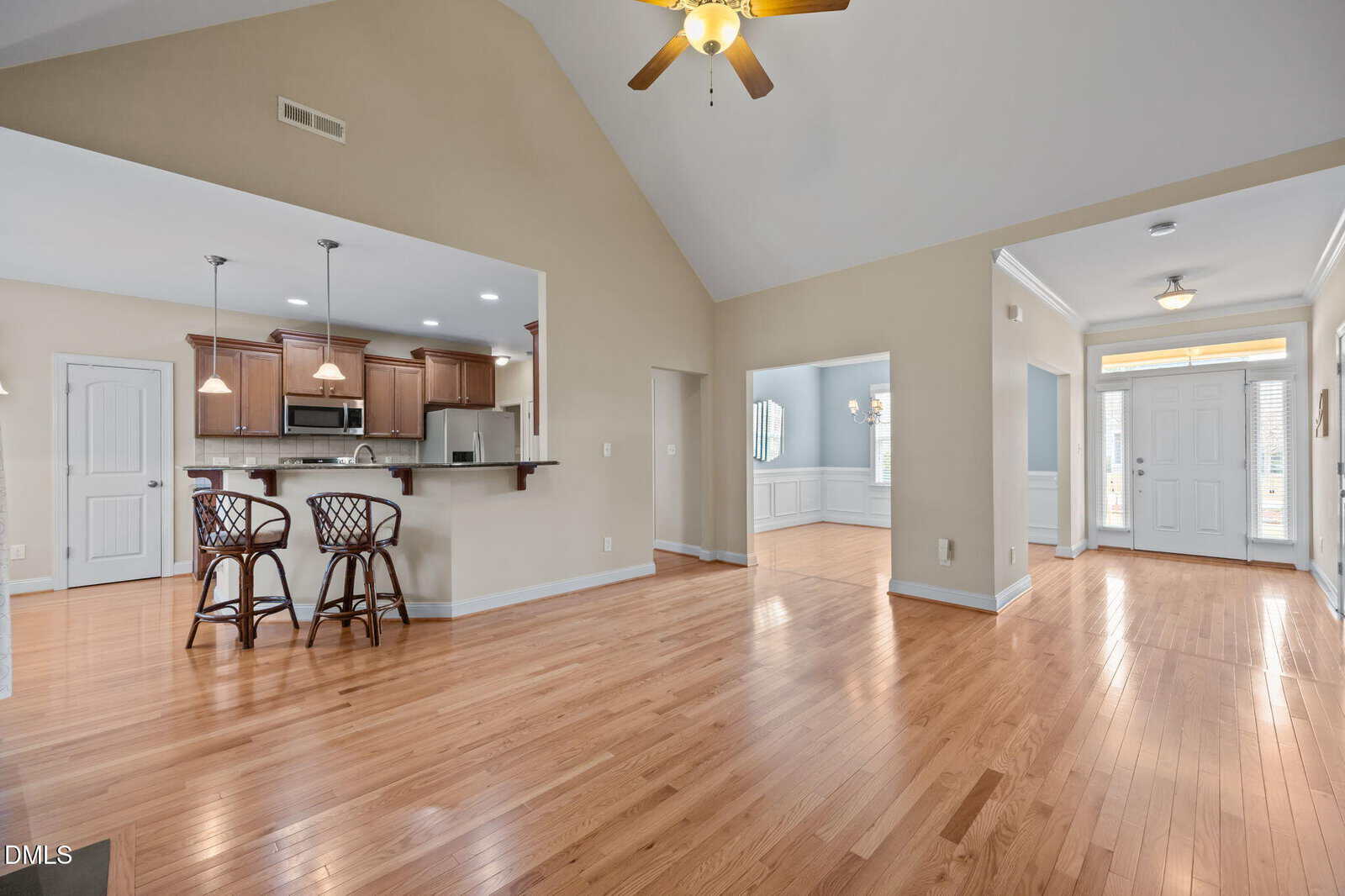 An open-concept living area with hardwood floors, vaulted ceilings, a ceiling fan, and a view of the kitchen with bar seating. Natural light enters through the windows by the front door and dining area.