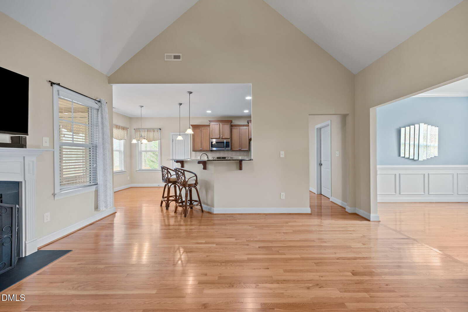 Open living area with hardwood floors, vaulted ceiling, fireplace, and view into a kitchen with bar seating. Natural light enters through multiple windows, creating a bright, spacious atmosphere.
