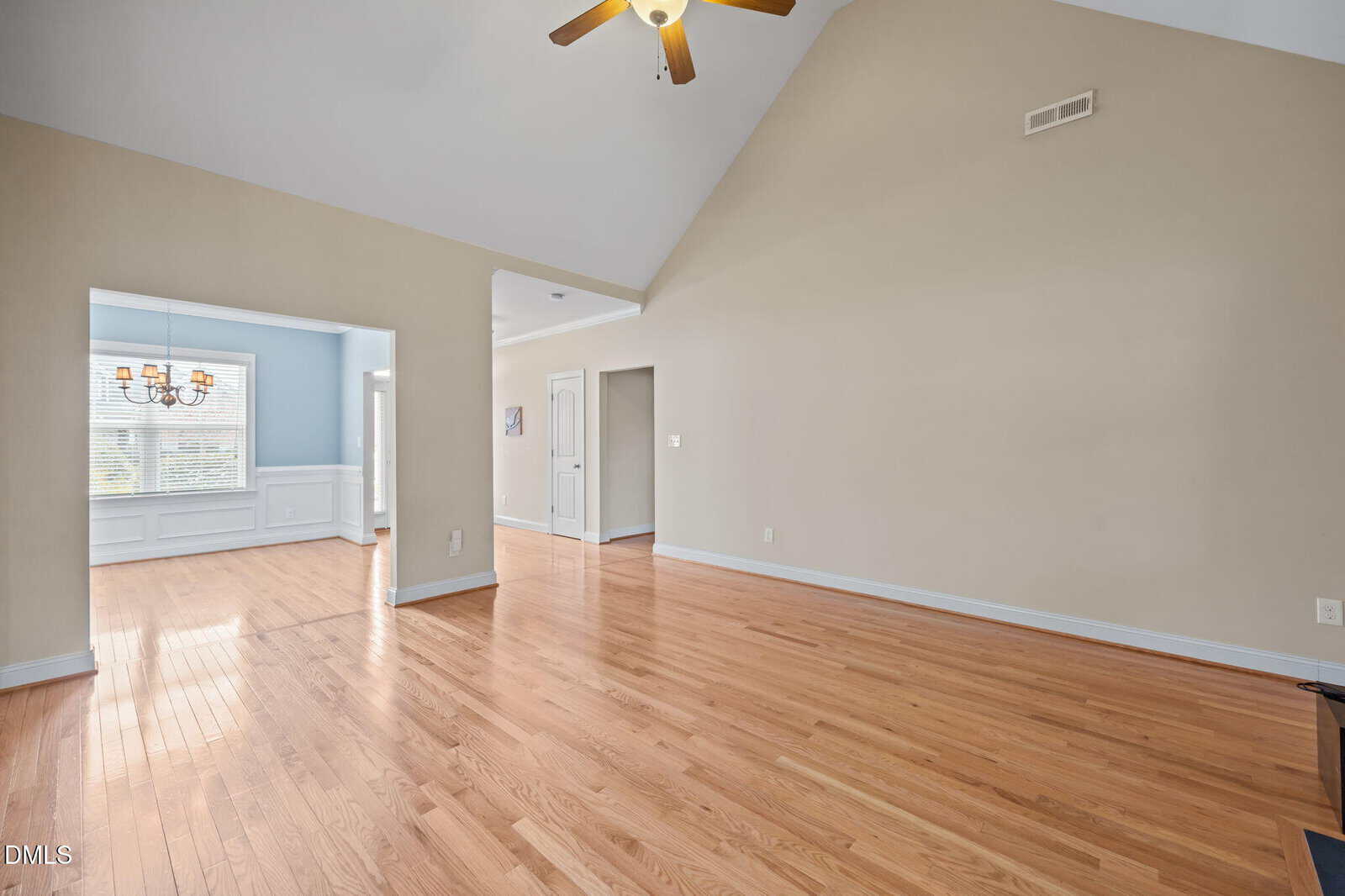 Spacious room with light wood floors, beige walls, vaulted ceiling, ceiling fan, and a view into a dining area with blue walls and a chandelier. Natural light fills the space.