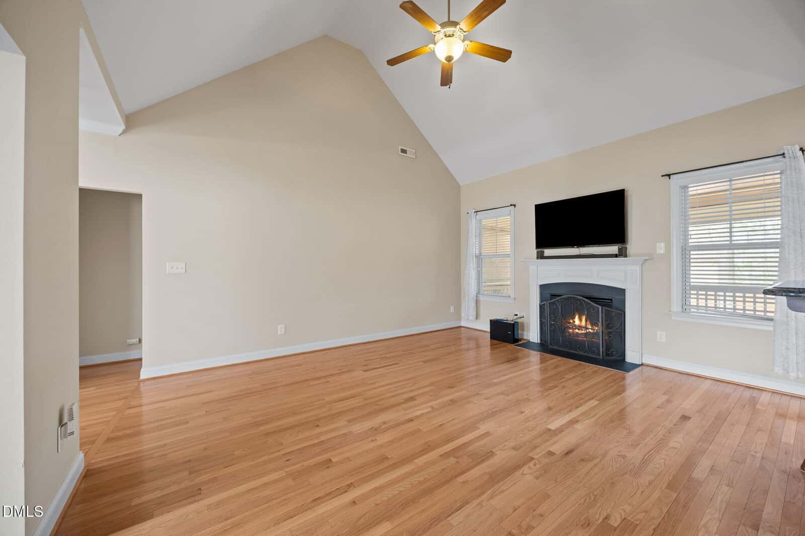 A spacious living room with light wood floors, beige walls, a vaulted ceiling with a ceiling fan, a fireplace with a TV mounted above it, and windows with white curtains allowing natural light in.