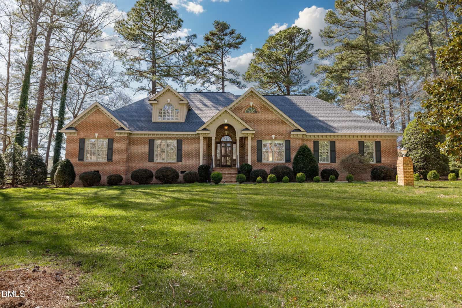 A single-story brick house with multiple gables, large windows, and a central arched doorway, set on a well-maintained lawn with trimmed bushes and tall trees in the background.