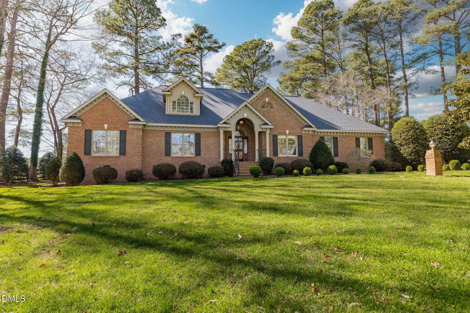 A single-story brick house with dormer windows sits on a large, neatly manicured lawn, surrounded by tall trees under a partly cloudy sky.