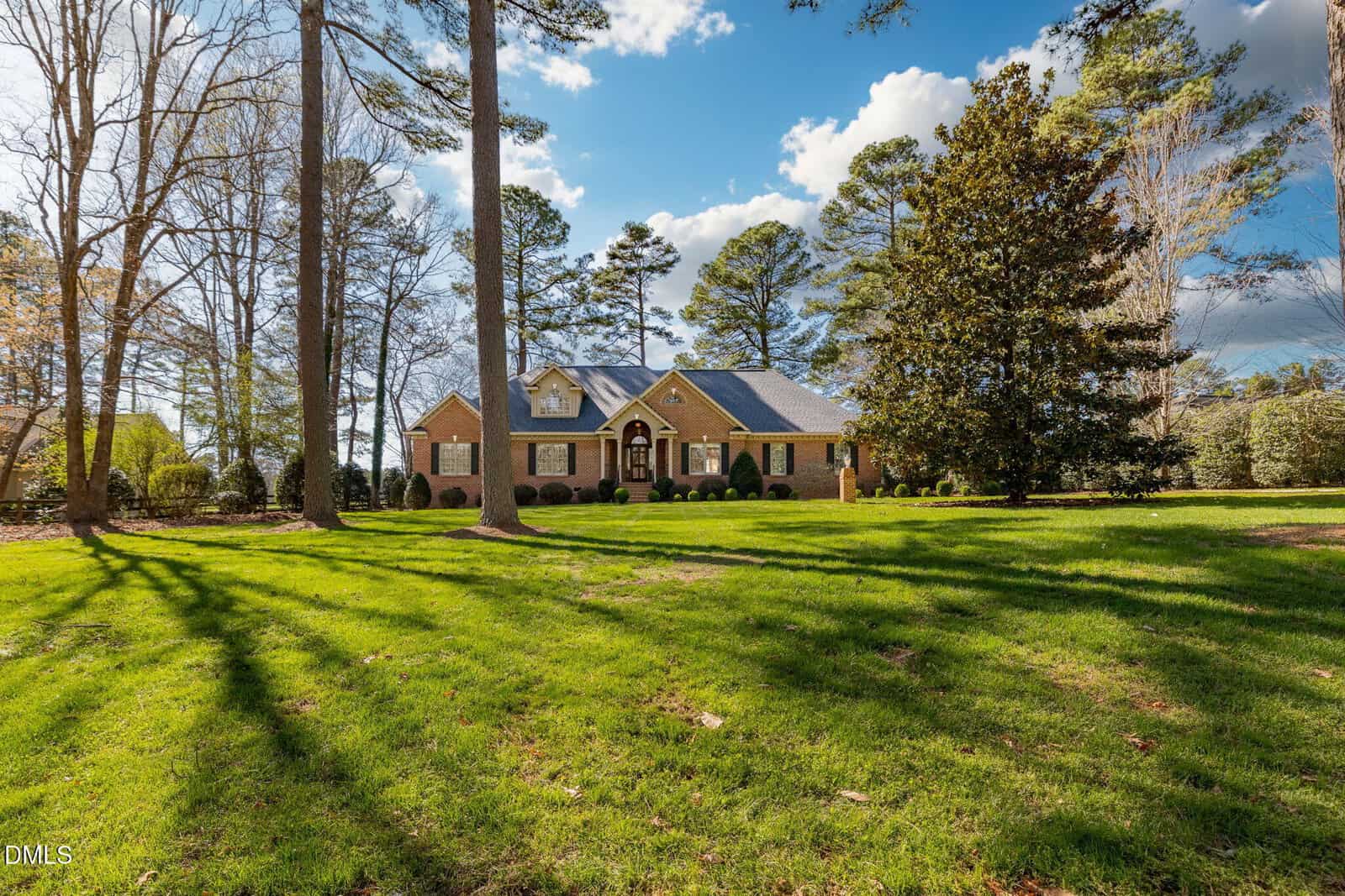 Brick house with black shutters and arched entry sits on a large, green lawn surrounded by tall trees. The sky is blue with scattered clouds, and shadows from the trees stretch across the grass.