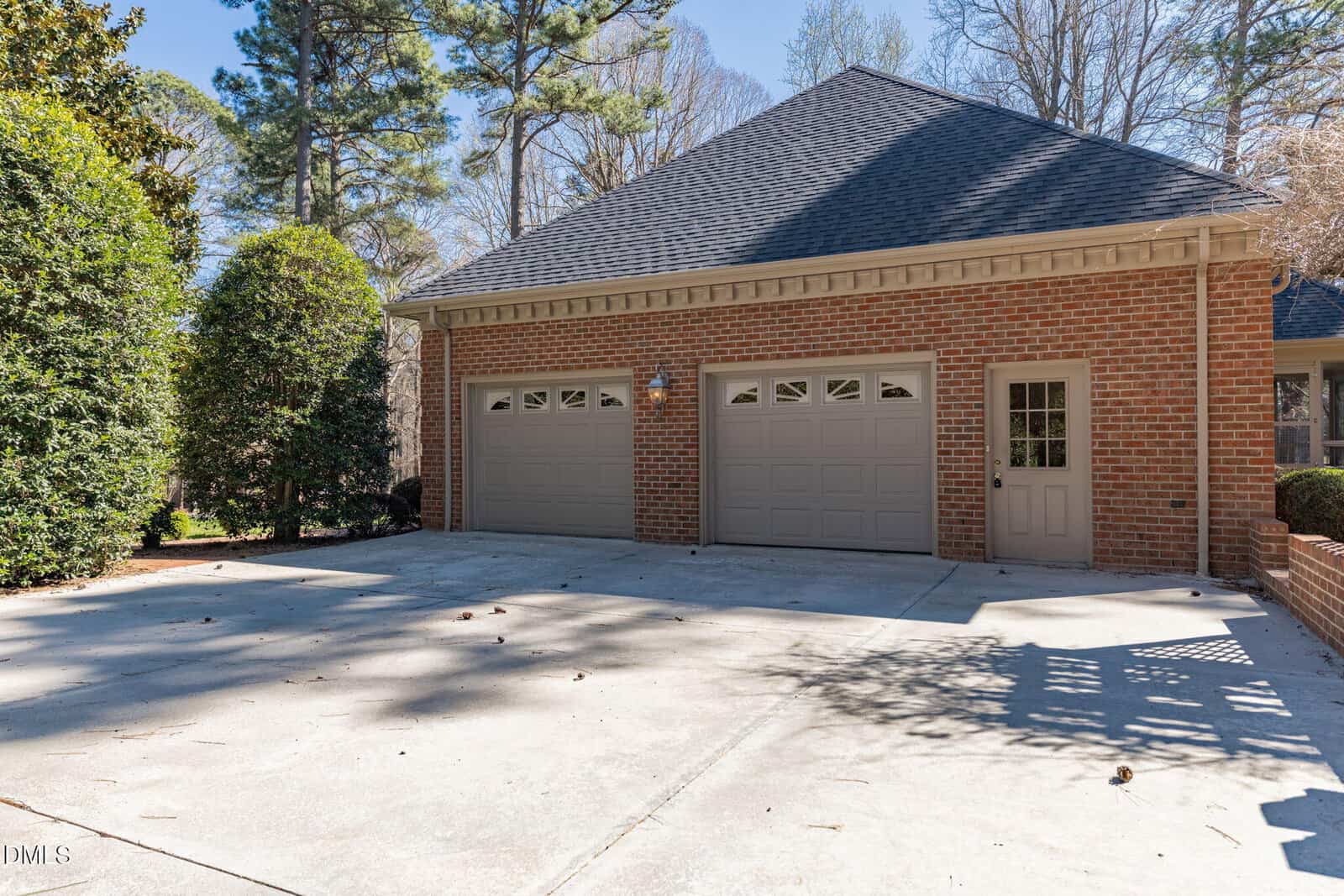 A brick house with two-car garage, beige doors, and a single side door. Wide concrete driveway in front, surrounded by tall trees and greenery under a clear blue sky.