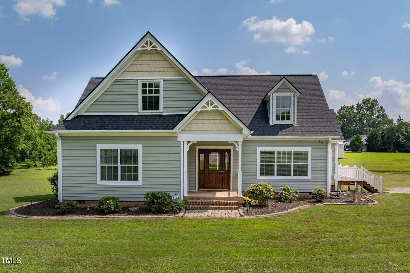 A light gray house with white trim, a dark roof, and a wooden front door sits on a neatly landscaped lawn with shrubs and trees in the background under a partly cloudy sky.
