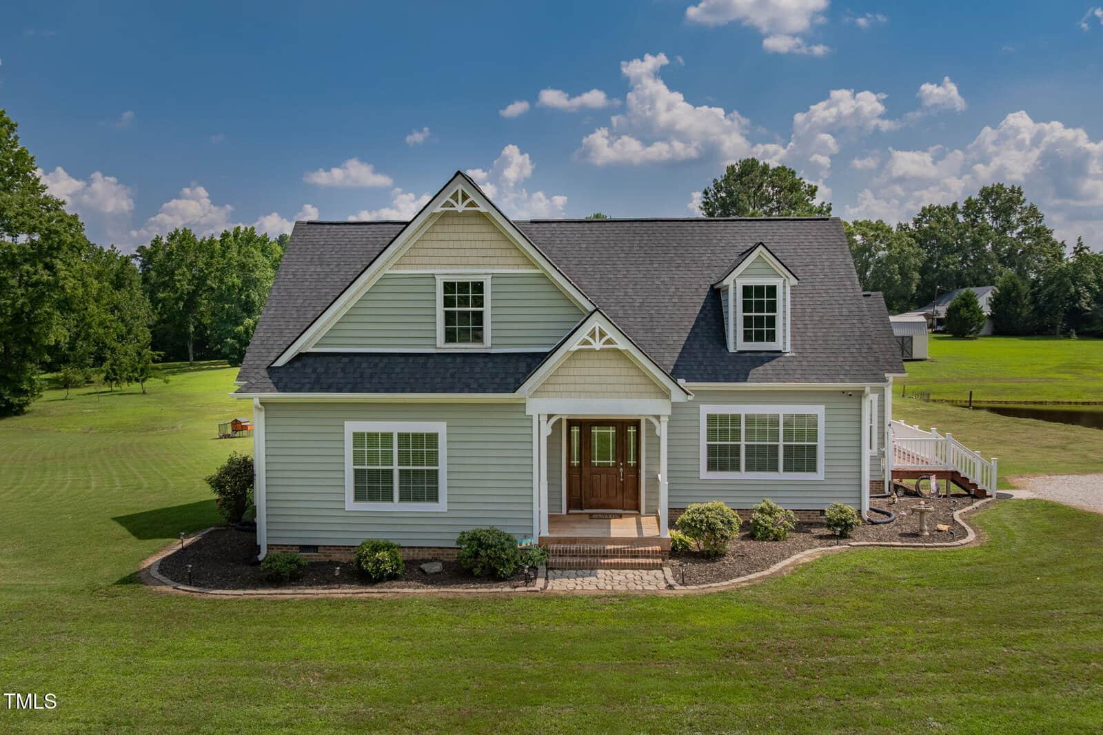 A two-story light gray house with white trim, a gabled roof, and a wooden front door sits on a large, well-maintained lawn with minimal landscaping. Trees and a partly cloudy sky are visible in the background.