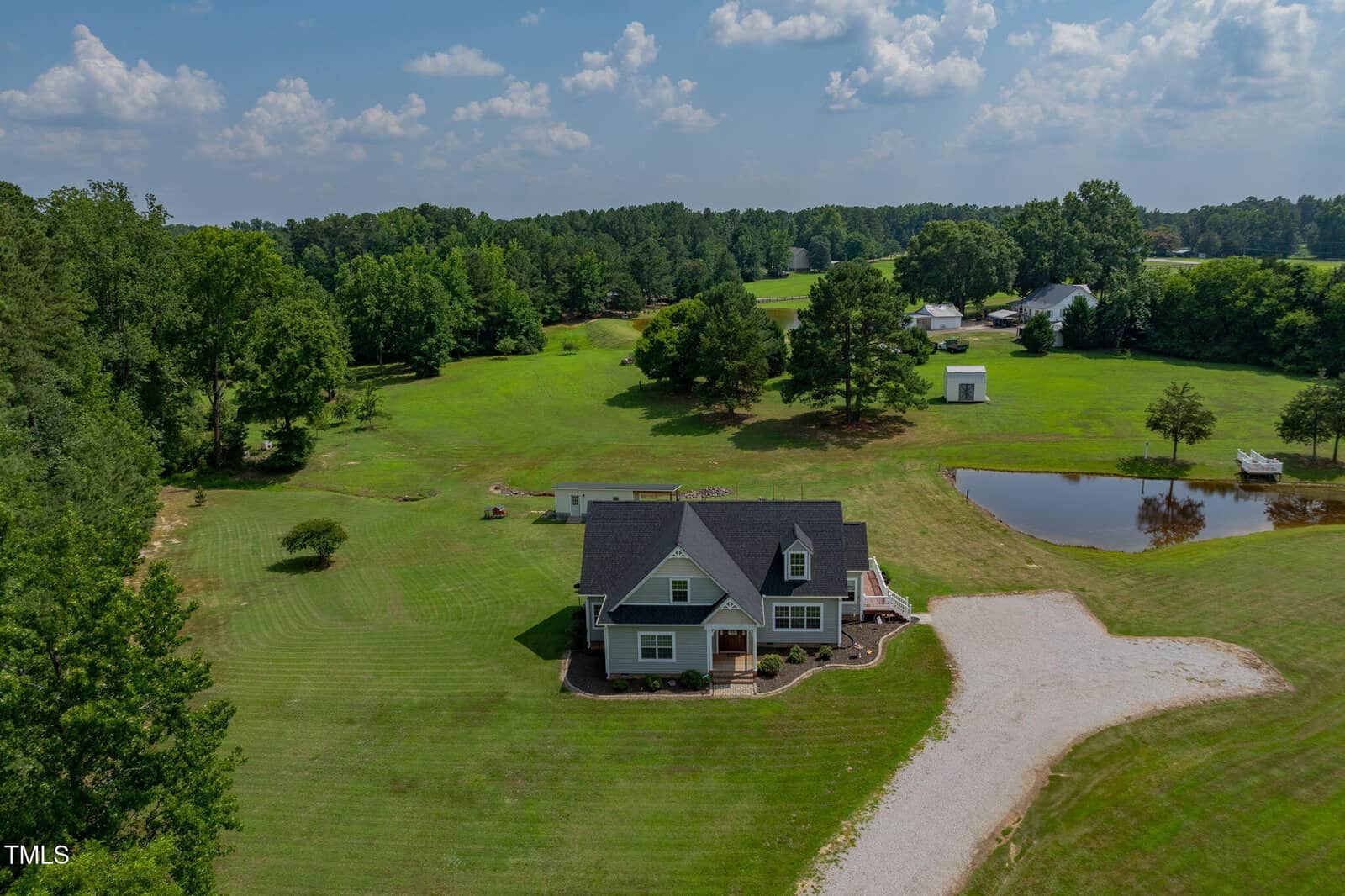 A single-family house with gray siding and a dark roof sits on a large, well-maintained grassy lot with a gravel driveway, a pond, scattered trees, and other buildings visible in the background.