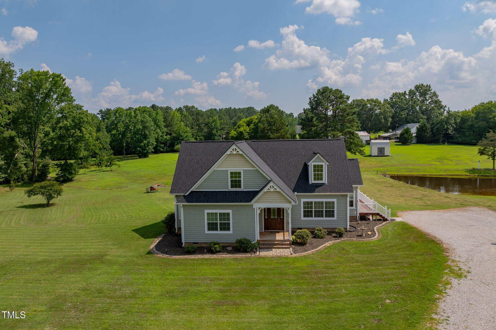 A gray, two-story house with dark roof sits on a large, neatly mowed lawn with trees, a gravel driveway, a small pond, and another structure visible in the background under a partly cloudy sky.
