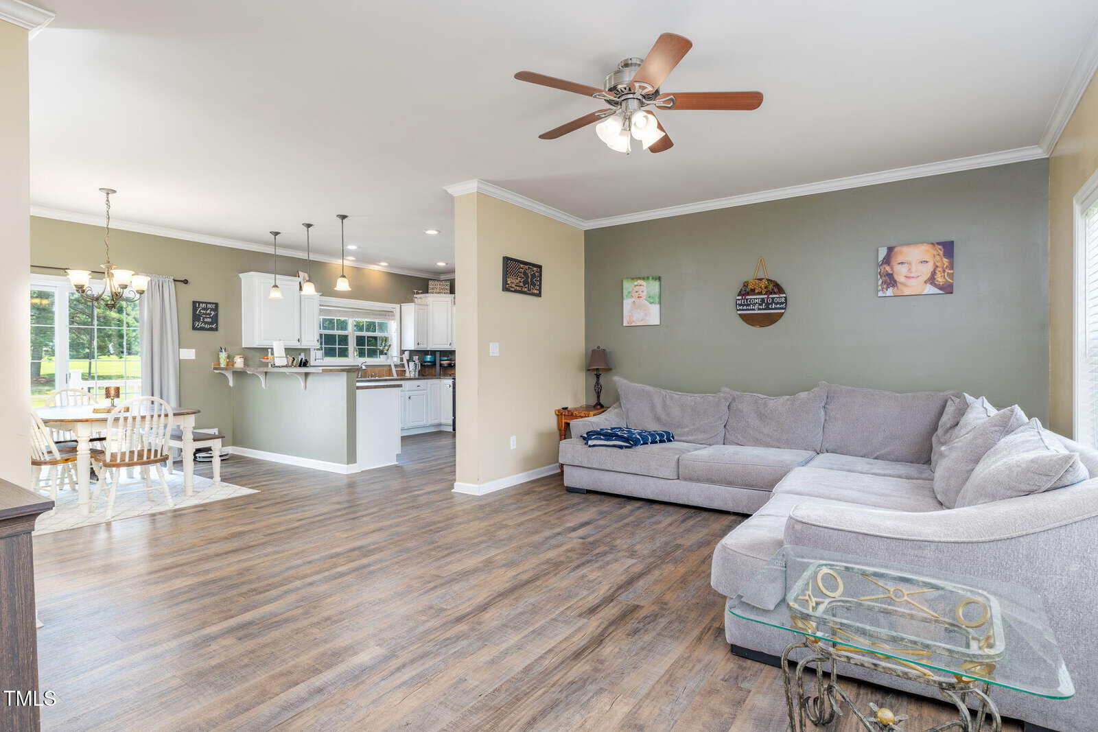 Spacious living room with a gray sectional sofa, wood flooring, ceiling fan, and wall art. Open layout connects to a dining area and white kitchen with pendant lighting. Large windows provide natural light.