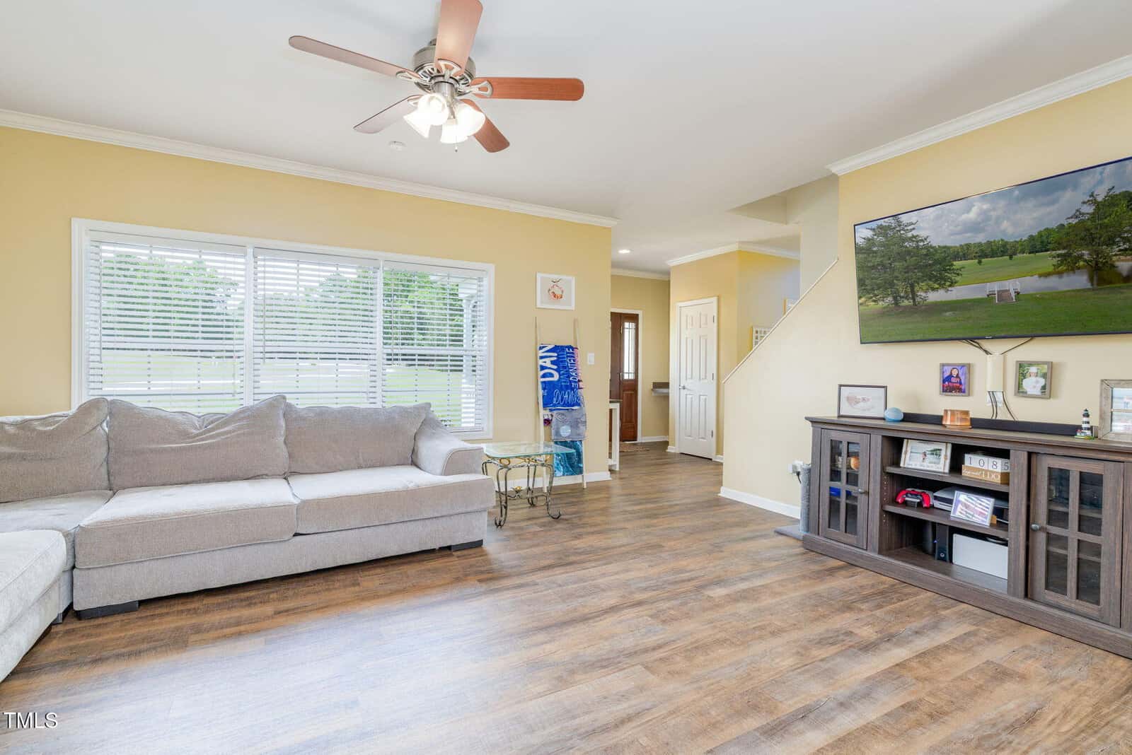 Bright living room with hardwood floors, beige sectional sofa, glass side table, wall-mounted TV, and large windows with blinds. A staircase and hallway are visible in the background.