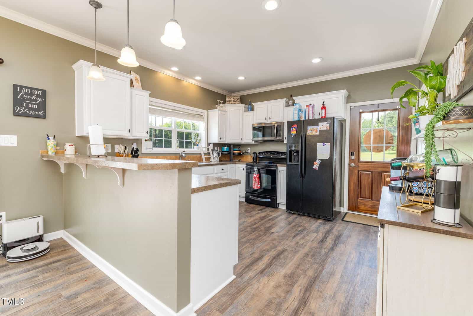 Modern kitchen with white cabinets, black appliances, wood floors, hanging lights, a large window, a coffee station, and a wooden door leading outside. A small robot vacuum is on the floor by the counter.