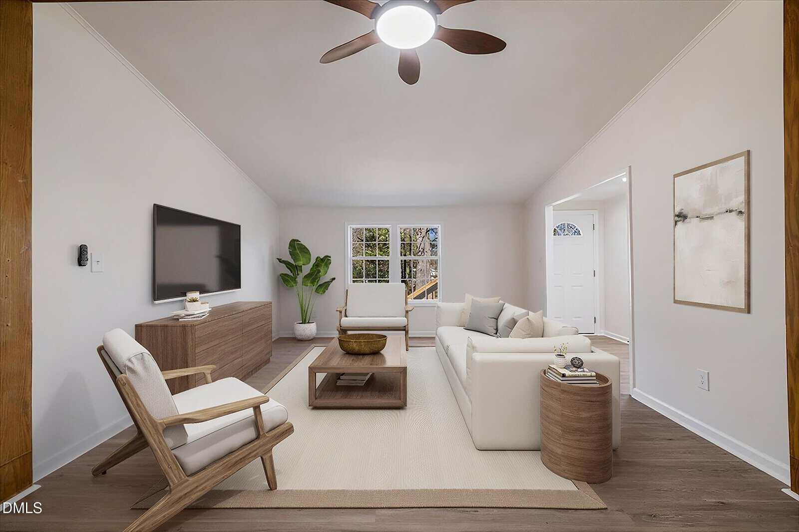 Modern living room with neutral decor, white sofas, wooden furniture, wall-mounted TV, large window, potted plant, and ceiling fan. Walls are light-colored, and floor is wood. Minimalist artwork hangs near the front door.