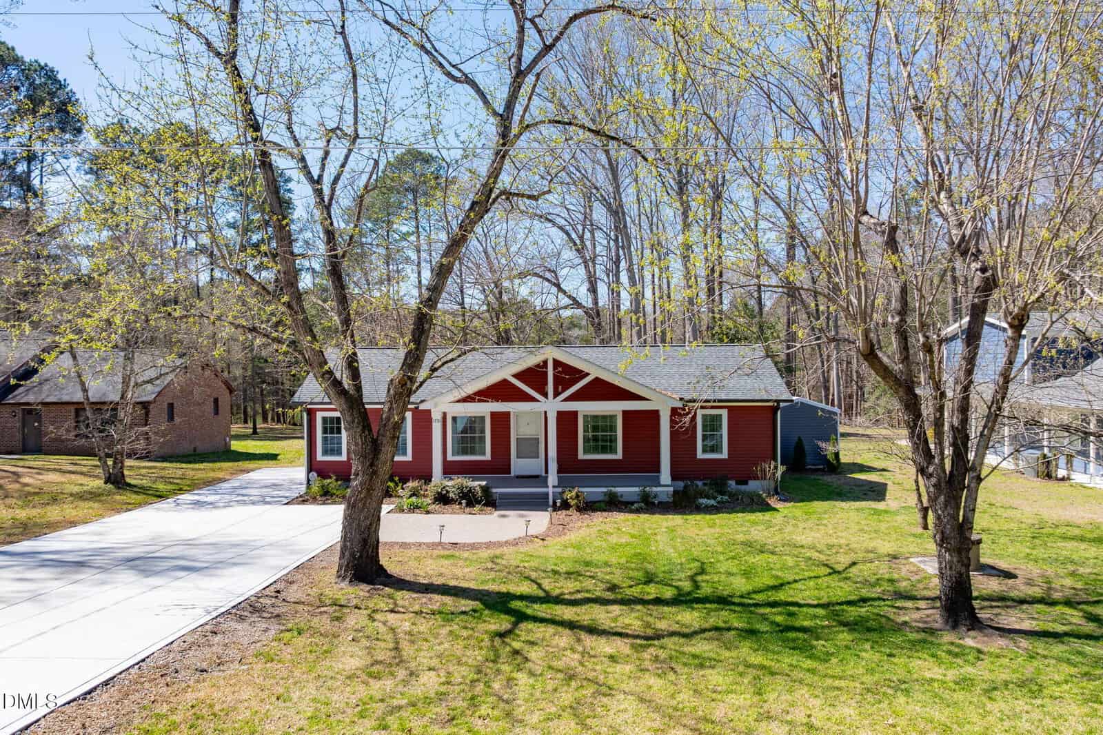 A single-story red house with white trim sits on a grassy lot with trees. There is a concrete driveway on the left, and the yard is partially shaded by large trees. Other houses are visible in the background.