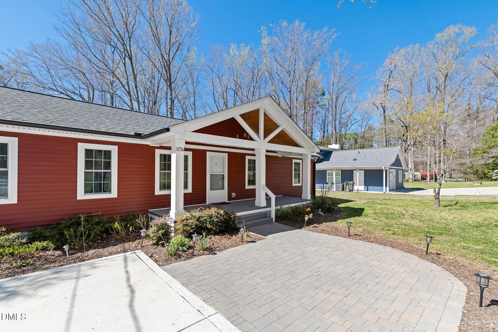 A red single-story house with white trim and a covered front porch, surrounded by shrubs and trees with no leaves. The yard features a paved walkway and driveway, and another house is visible in the background.