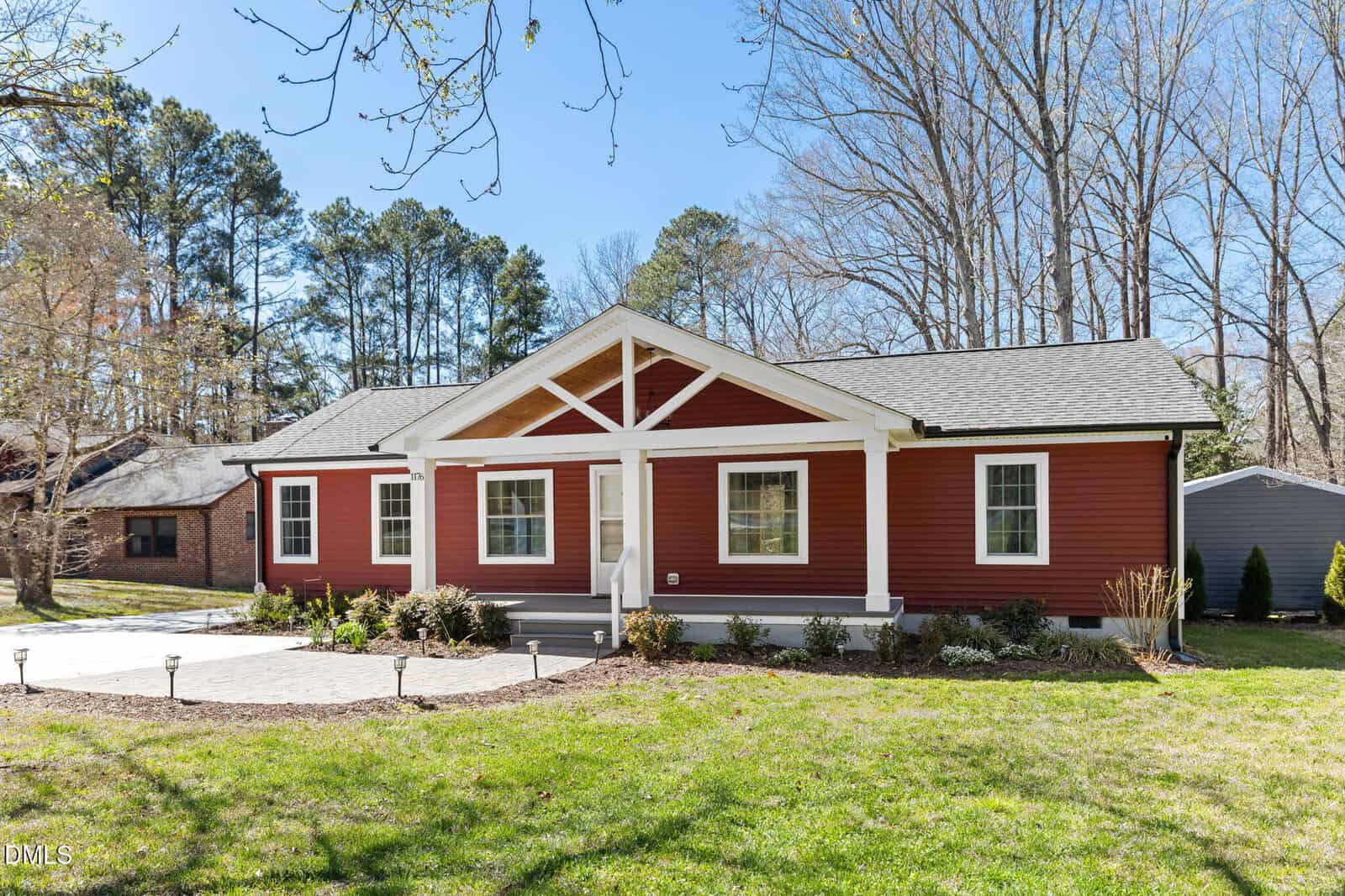 A single-story red house with white trim, a covered front porch, and three large windows sits on a grassy lawn with a paved walkway. Mature trees and neighboring houses are visible in the background.