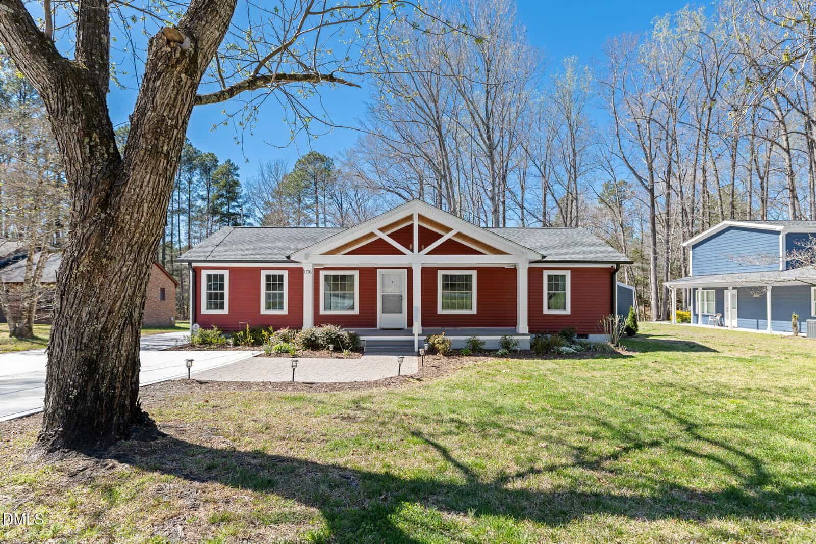 A single-story red house with white trim and a covered porch sits on a grassy lawn with a large tree in front. The house has multiple windows, a concrete driveway, and is surrounded by other homes and tall trees.
