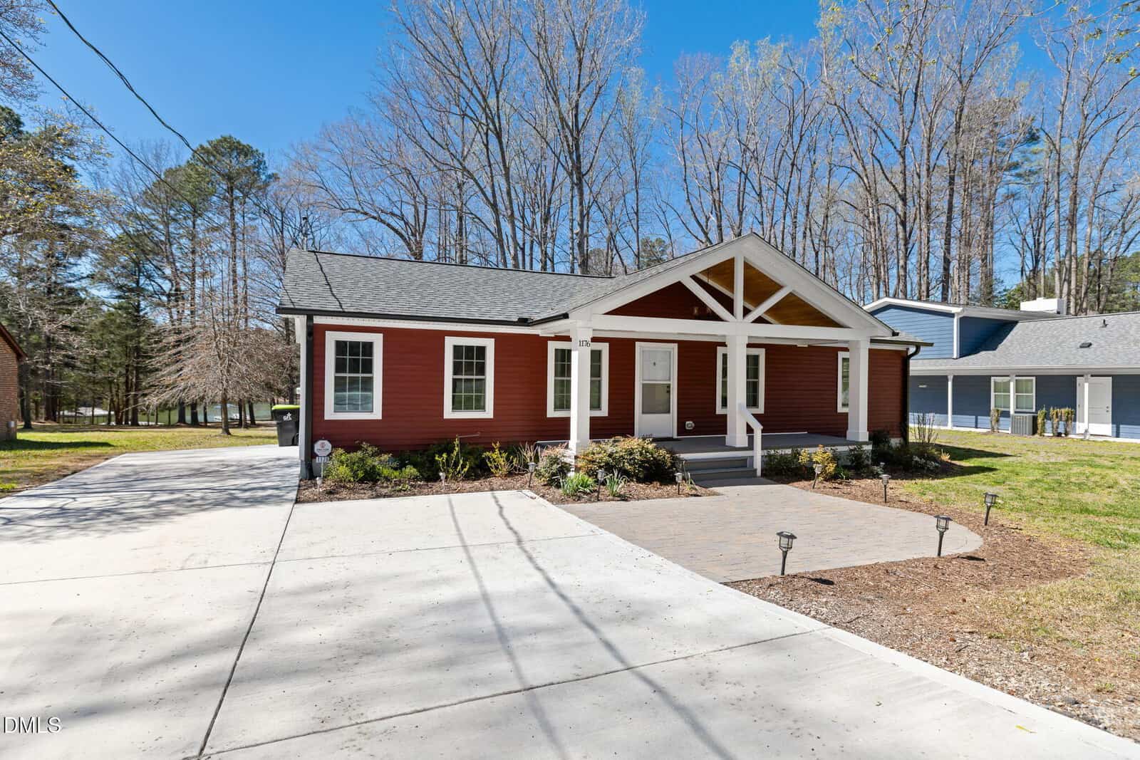 A single-story red house with white trim, a covered front porch with wooden beams, a paved driveway, and a small landscaped area, set against a backdrop of tall trees and clear blue sky.