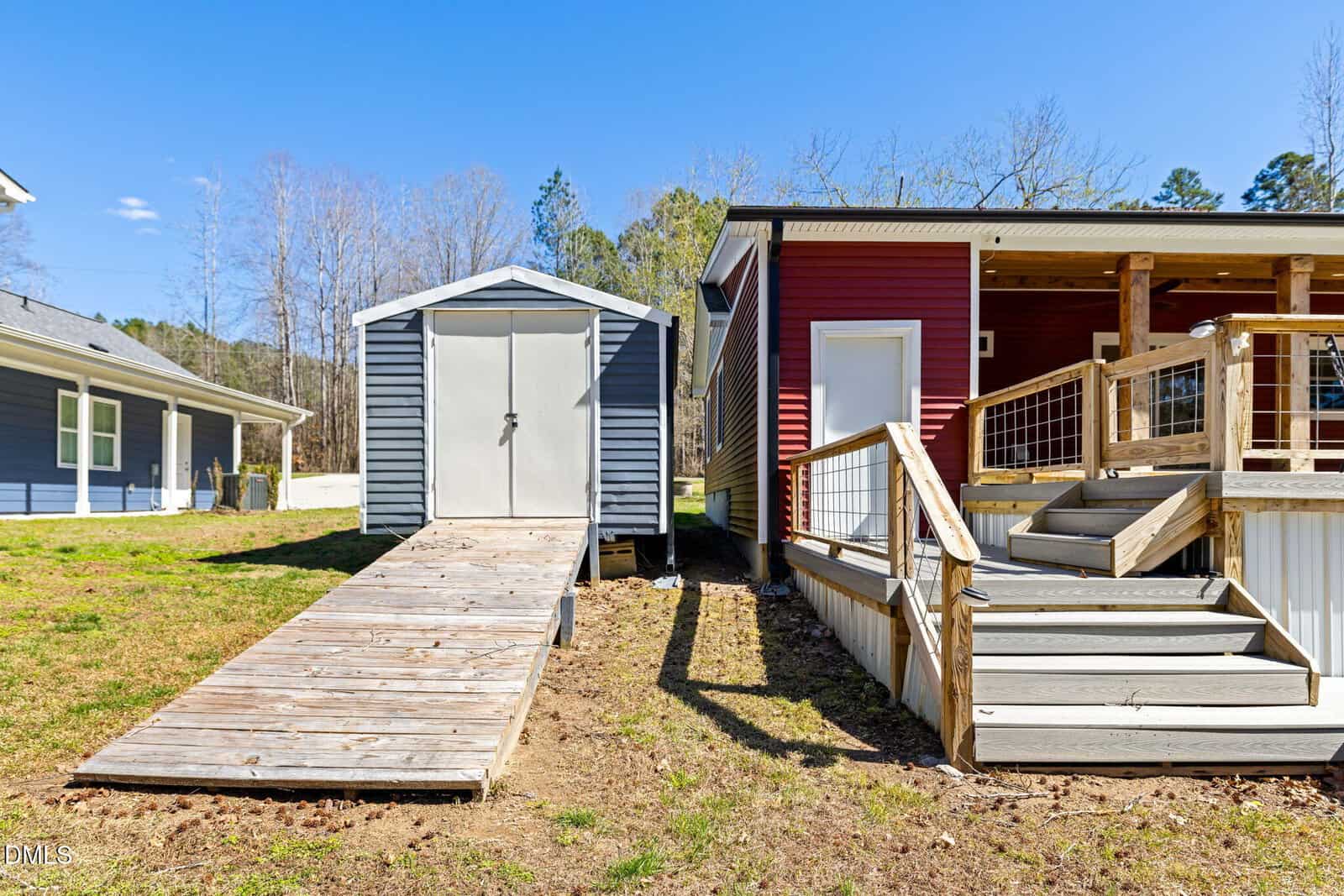 A gray storage shed with a wooden ramp stands next to a red house with a raised porch and stairs. Both structures are set on a grassy lawn under a clear blue sky.