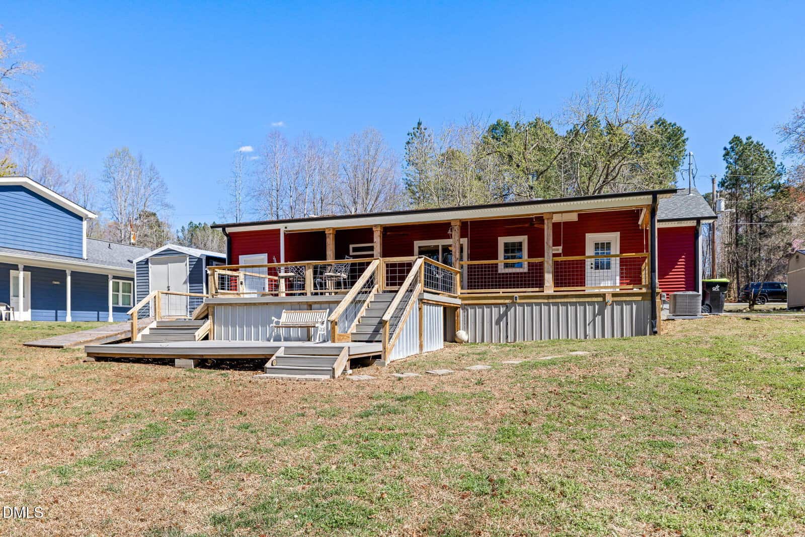 A red single-story house with a spacious wooden deck, railings, and stairs leading to a grassy yard. Other houses and tall trees are visible in the background under a clear blue sky.