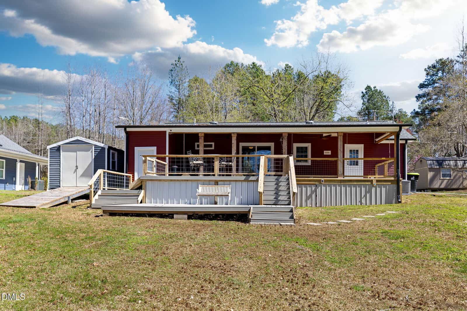 A red house with a covered front porch, wooden railing, and steps leading to a grassy yard. A shed and trees are visible in the background under a partly cloudy sky.