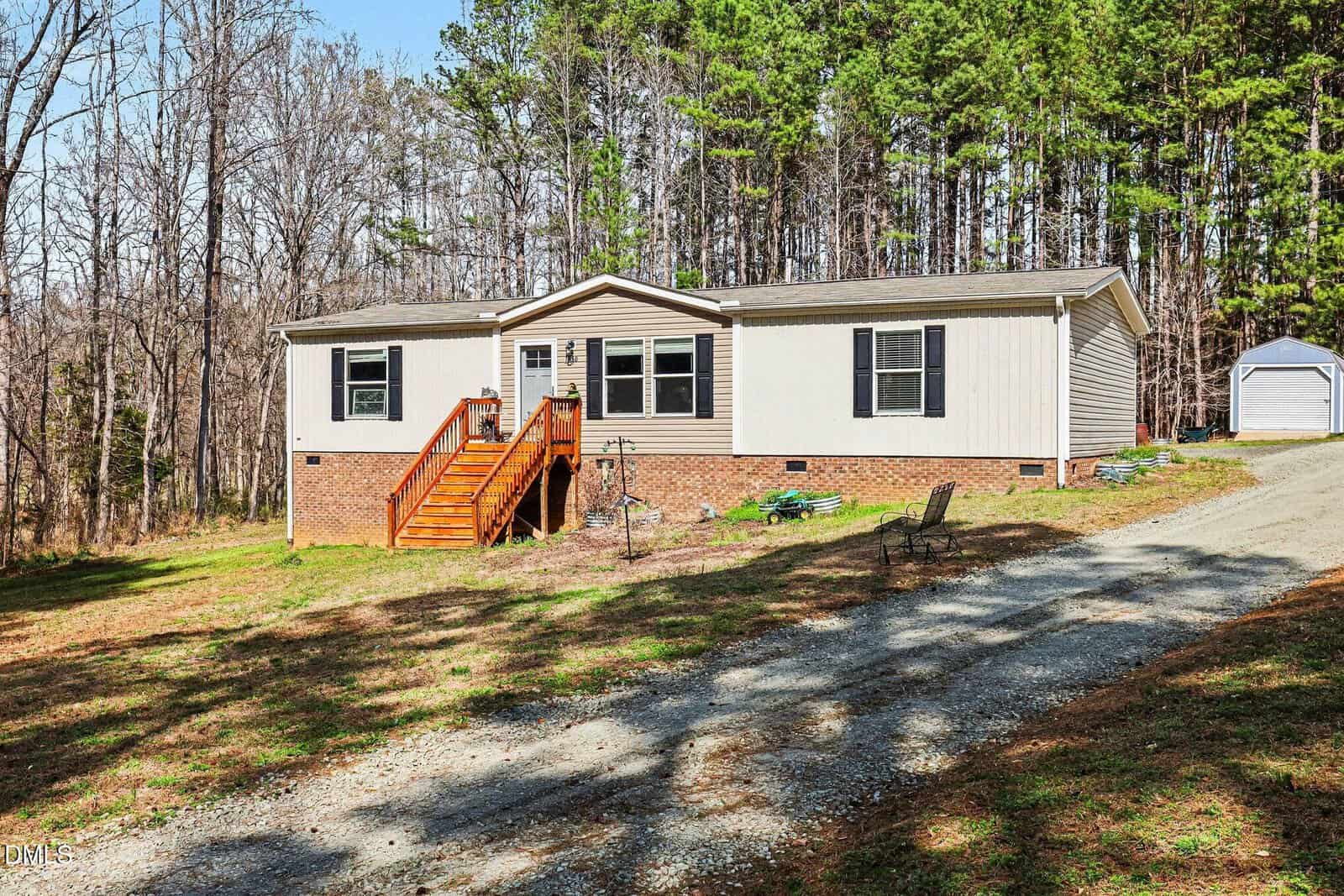 A single-story house with beige siding, black shutters, and a brick foundation. Wooden steps lead to the front door. The house sits near a wooded area, with a gravel driveway and a small shed in the background.