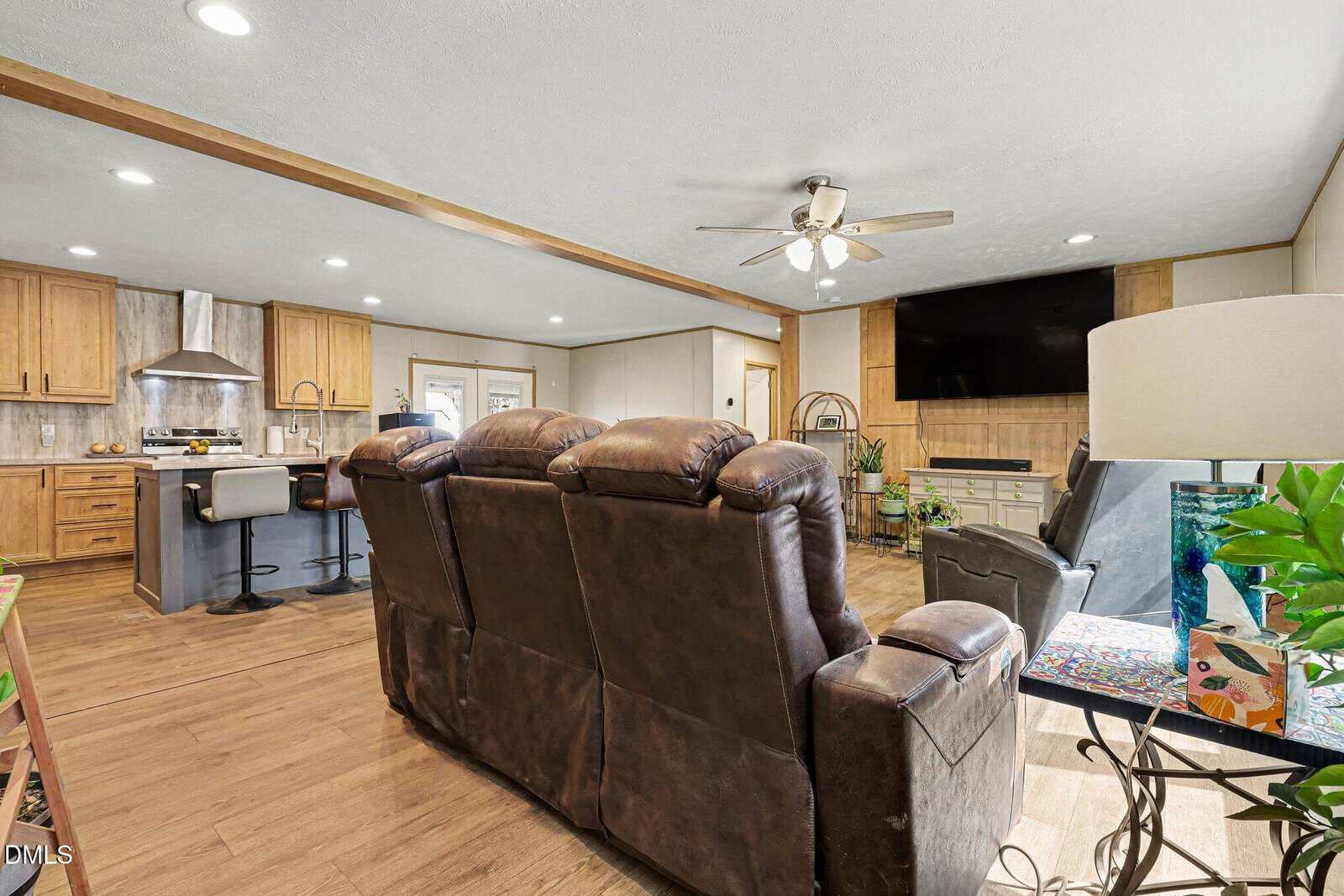 A living room with three brown recliners facing a mounted TV above a fireplace. The open layout leads to a kitchen with wooden cabinets, an island, and stainless steel appliances. Plants and a lamp are placed near the seating area.