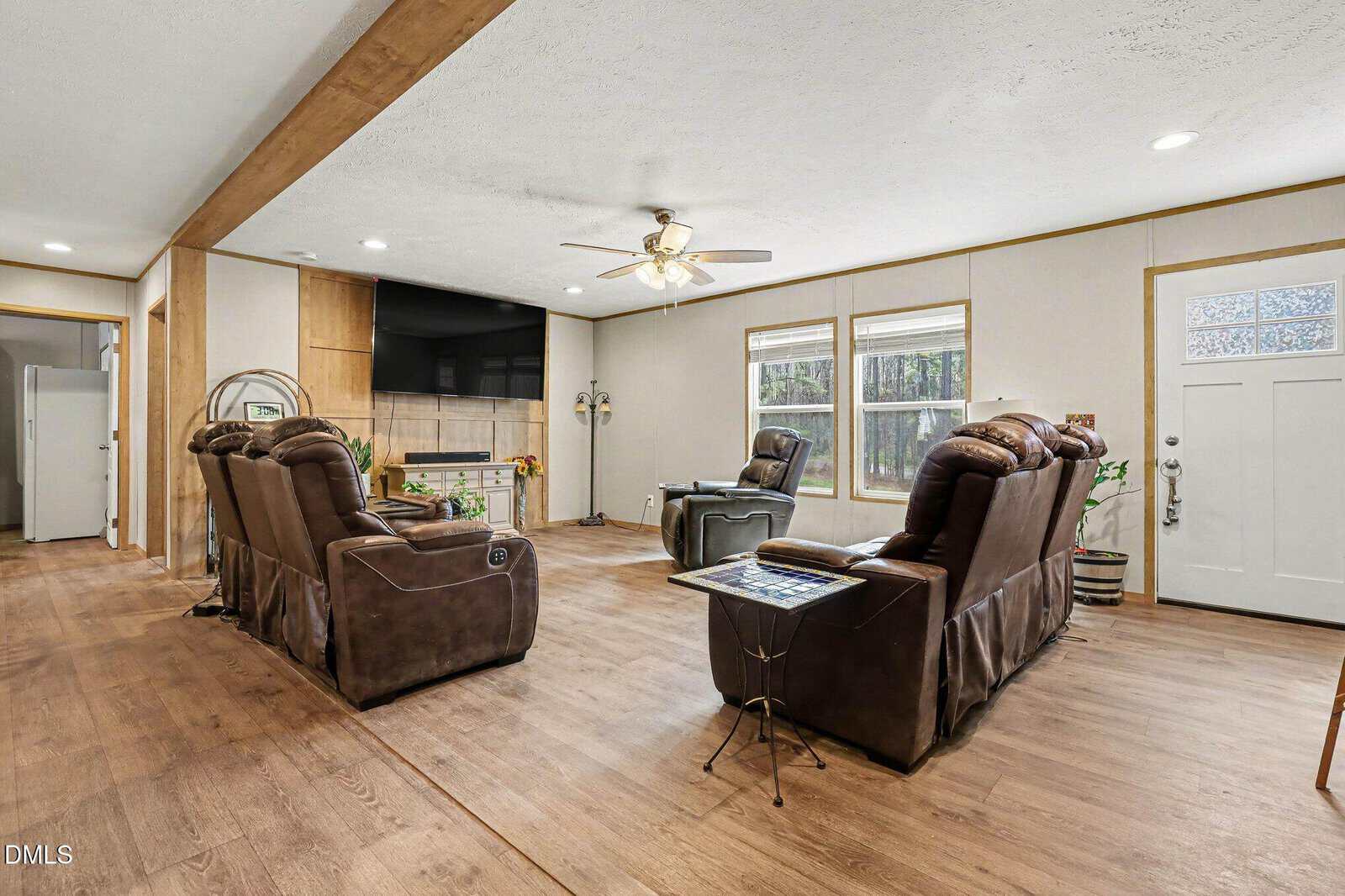 A living room with wood flooring, two brown recliner sofas, a large TV on a tiled accent wall, a ceiling fan with lights, and windows letting in natural light. The front door is visible on the right.
