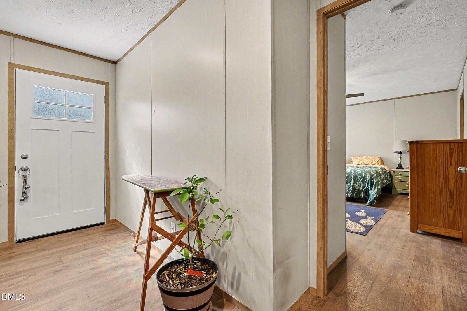A hallway with wood flooring features a white front door with a window, a wooden folding table, and a potted plant. An open doorway leads to a bedroom with a green dresser, bed, and nightstand.