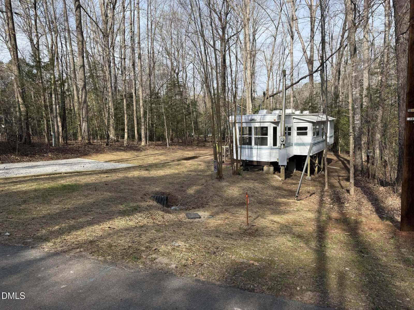 A small white trailer is elevated on stilts in a clearing surrounded by bare trees. There is a gravel parking area on the left and a driveway in the foreground, with patchy grass and scattered leaves on the ground.
