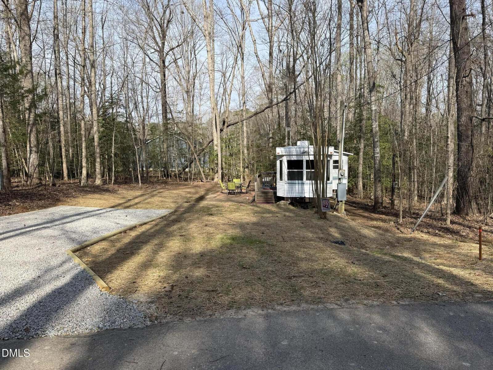 A small white travel trailer is parked in a wooded area with leafless trees. There is a gravel parking spot on the left and a few chairs and a barrel near the trailer. The ground is mostly dirt and dry grass.