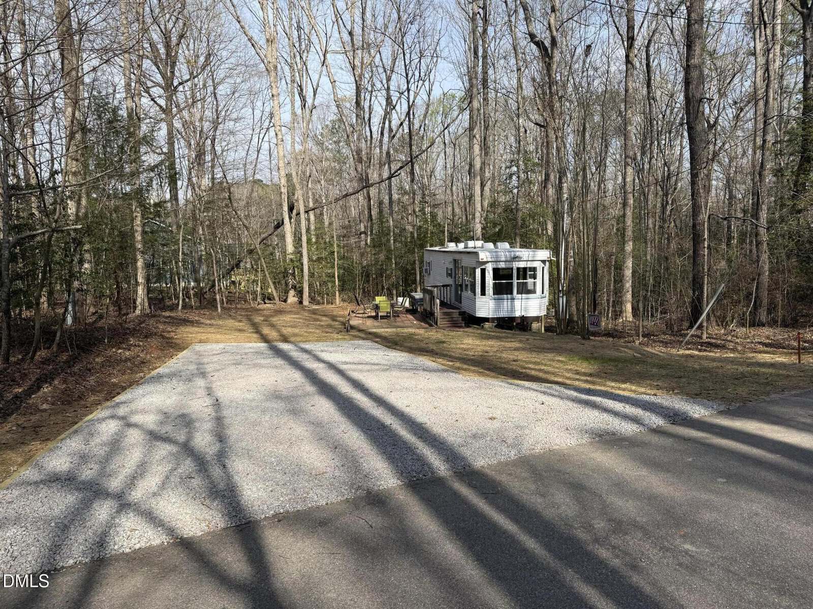 A gravel parking area is next to a wooded lot with bare trees. A small white RV trailer is parked near the trees, with two chairs and a picnic table nearby. The scene is lit by daylight.