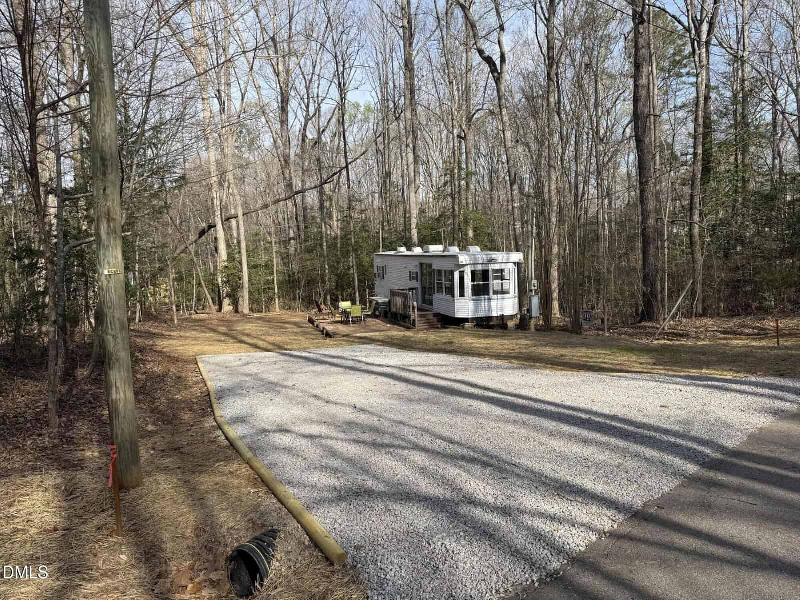 A small white trailer is parked in a clearing surrounded by tall, leafless trees. There is a gravel parking area in the foreground and some patio furniture next to the trailer. The setting appears peaceful and wooded.